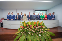 A imagem mostra um grupo de pessoas perfiladas em pé, sobre um palco elevado em um auditório. Ao centro e à frente, há um grande arranjo de flores tropicais coloridas que compõem a decoração. As pessoas, em sua maioria de terno ou trajes sociais, parecem participar de uma solenidade formal. No fundo, há uma parede branca com projetor, reforçando o ambiente institucional da cerimônia.