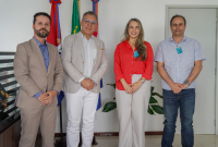 Na imagem, quatro pessoas estão em pé, lado a lado, posando para a foto em um ambiente interno. Atrás delas, há bandeiras e uma parede branca. Duas pessoas estão de terno, uma veste camisa social clara e calça jeans, e outra usa blusa vermelha e calça clara. Todos estão sorrindo.