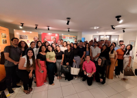 A imagem mostra um grupo grande de pessoas reunidas em um espaço interno, semelhante a uma exposição. Elas estão de pé, sorrindo para a foto, em frente a painéis coloridos com textos e imagens. O ambiente é iluminado por spots no teto e tem piso claro. É uma visita em grupo ao Memorial Pontes de Miranda do TRT-AL.