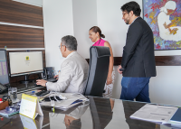 Na imagem, três pessoas estão reunidas em um ambiente de trabalho, diante de uma mesa com computador. Um homem, sentado, interage com a tela enquanto os outros dois, de pé, observam atentamente. O clima é descontraído, evidenciado pelos sorrisos, sugerindo uma apresentação ou demonstração. Ao fundo, destaca-se uma pintura colorida, que contrasta com o mobiliário moderno do escritório.