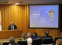 Homem de terno fala em um púlpito com microfone, em auditório. À direita, há um telão projetando a apresentação do “Corisco Chat JT”, com a imagem de um mascote em forma de robô vestido de cangaceiro. Pessoas estão sentadas assistindo à apresentação.