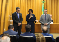Homem de paletó preto à esquerda, ao centro, mulher de calça preta e blusa preta, à direita, homem de paletó cinza. Todos estão em pé. A mulher está com um microfone na mão direita