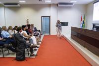 Na foto, uma mulher em pé apresenta-se com microfone em mãos diante de um grupo de pessoas sentadas, em ambiente de auditório. Ela está posicionada sobre um tapete vermelho, ao lado de uma mesa de autoridades e de duas bandeiras hasteadas. Os presentes acompanham atentos, alguns fazendo anotações e outros utilizando laptops. A cena transmite clima de palestra ou exposição institucional.