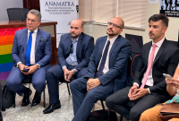 quatro homens sentados de terno com uma bandeira LGBTQIA+ ao fundo