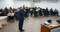 Há um homem de pé com um microfone na maão. Ele olha para frente . Na sala há várias pessoas sentadas. 