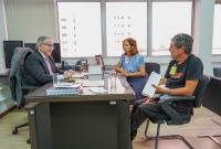 Três pessoa sentada ao redor de uma mesa, dois homens e uma mulher; há um homem de terno escuro que aparentemente está sentado do lado principal atendo o outro homem a a mulher, que vestem roupas informais