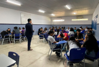 Há uma sala de aula com várias jovens. Eles estão sentados em círculos em bancas escolares. No centro da sala há um homem de pé. Ele está de terno escuro.