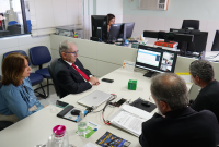A foto mostra uma reunião em um ambiente de escritório. Quatro pessoas estão sentadas ao redor de uma mesa, voltadas para um monitor de computador onde ocorre uma videoconferência. Entre elas, há um homem de terno e gravata vermelha, uma mulher de blusa azul, e dois homens de terno escuro. Na mesa, estão documentos, um notebook, garrafa de água, copo plástico, álcool em gel, celular, além de alguns papéis e folders institucionais. Ao fundo, uma servidora trabalha em outro computador. 