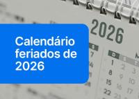 Foto cinza da página do calendário 2026 ao fundo; em destaque à esquerda, uma tarja azul com a frase Calendário feriados de 2026 escrita em letras brancas