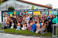 Varias pessoas, homens e mulheres, de roupas coloridas sentada e em pé em frente a um muro multicolorido