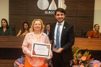 Mulher idosa à esquerda, de saia laranja e blusa rosa recebendo um diploma de uma advogado alto de paletó preto e gravata azul à direita. Ambos estão em pé ollhando para o fotógrafo
