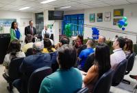 A foto mostra um auditório do Tribunal Regional do Trabalho da 19ª Região durante a abertura de um evento. Um grupo de pessoas está sentado, atento à fala de um dos oradores, que aparece ao centro segurando papéis. Ao fundo, há balões verdes e azuis e uma mesa com lanche, compondo uma decoração festiva. O ambiente é iluminado e transmite clima de acolhimento e engajamento institucional.