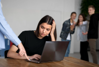 A imagem mostra uma mulher sentada à mesa, usando um notebook, com expressão triste e preocupada. Ela apoia uma das mãos na cabeça enquanto uma pessoa ao lado aponta algo na tela do computador. Ao fundo, três pessoas estão em pé, rindo e apontando em sua direção, sugerindo uma situação de constrangimento ou assédio no ambiente de trabalho.