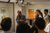 Um grupo de estudantes observa um homem que fala e gesticula durante uma visita guiada a um espaço expositivo. Ao fundo, há vitrines com documentos e painéis coloridos com textos e fotos históricos. O ambiente é bem iluminado, com paredes claras e iluminação direcionada para as peças expostas.