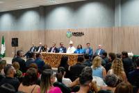 Há uma cerimônia formal em um auditório da UNCISAL (Universidade Estadual de Ciências da Saúde de Alagoas). No palco, há uma mesa com nove pessoas sentadas, entre elas homens e mulheres, com trajes formais. Ao fundo, o logotipo da universidade está fixado na parede. À frente do palco, o público assiste atentamente ao evento, com várias pessoas sentadas em fileiras. O ambiente é bem iluminado e transmite um clima solene.