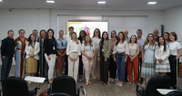 A imagem mostra um grupo de pessoas posando para uma foto em um ambiente interno, possivelmente uma sala de treinamento ou auditório. Elas estão organizadas em uma única fileira, sorrindo para a câmera. Ao fundo, há um telão ligado exibindo uma apresentação com texto e imagem, sugerindo que o grupo participou de uma atividade ou evento institucional. O ambiente é bem iluminado, com piso de madeira e cadeiras de cor escura dispostas à frente do grupo.