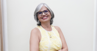 A imagem mostra uma mulher de cabelos grisalhos curtos, usando óculos e sorrindo. Ela veste um vestido amarelo de renda clara e está posando de braços cruzados diante de um fundo neutro, transmitindo simpatia e confiança.