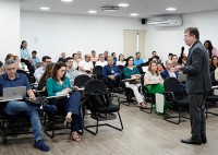 A imagem mostra um homem de terno falando ao microfone diante de um auditório com várias pessoas sentadas. Os participantes, que parecem servidores ou profissionais, acompanham atentamente a apresentação, alguns fazendo anotações ou utilizando notebooks. O ambiente é uma sala ampla, bem iluminada, com piso de madeira, cadeiras pretas e ar-condicionado. Ao fundo, observa-se uma porta com persiana e mais pessoas assistindo à palestra. A cena transmite um clima de concentração e aprendizado.