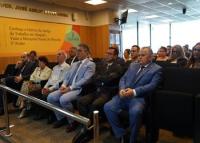 A imagem mostra um grupo de pessoas sentadas em um auditório, acompanhando atentamente um evento institucional. Na primeira fileira, estão homens e mulheres trajando roupas formais, incluindo ternos e vestidos, o que sugere a presença de autoridades ou convidados de destaque. O ambiente é bem iluminado e possui um painel na parede com a inscrição “Conheça a história da Justiça do Trabalho em Alagoas. Visite o Memorial Pontes de Miranda. 3º andar”. 