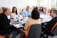Várias pessoas reunidas. Elas estão sentadas e ouvem um homem de terno escuro falar