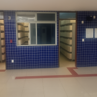 A imagem mostra um corredor interno com piso branco brilhante e faixas táteis vermelhas no chão, indicativas de acessibilidade. As paredes são revestidas por pequenos azulejos azuis com rejunte branco, formando um visual organizado e limpo. Há três portas abertas que revelam salas com estantes cheias de caixas de arquivo, sugerindo tratar-se de um arquivo ou setor de armazenamento de documentos. Na parede à direita, há um extintor de incêndio e, acima dele, um ponto de iluminação. 