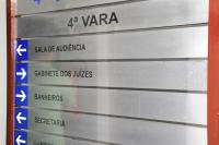 Placa de metal com moldura em madeira, há letras pretas escrito em destaque 4ª Vara.. Há setas indicativas.