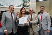 A foto mostra um momento de homenagem durante uma solenidade. Quatro pessoas posam sorridentes para a câmera em um ambiente formal, com bandeiras e painel institucional ao fundo. Ao centro, uma mulher segura um certificado de Moção de Aplausos, enquanto é acompanhada por três homens, todos trajando ternos. O clima é de celebração e reconhecimento, com destaque para a entrega do diploma que simboliza a homenagem prestada.