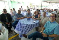Várias pessoas reunidas em voltade uma mesa que tem uma toalha de xadres azul. Aofundo há outras mesas também ocupadas por diversas pessoas.