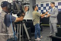 Há um homem de terno cinza claro sendo entrevistado, por um repórter qeu veste camisa verde e calça jeans. Como também aparece o cinegrafista com a filmadora.