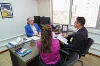 A imagem mostra três pessoas reunidas em um escritório. Um homem de terno azul está sentado atrás da mesa, conversando com um homem de terno escuro e uma mulher de blusa roxa, sentados à frente dele. Sobre a mesa há documentos, cadernos, celular, calculadora e outros itens de trabalho. Ao fundo, há janelas amplas com vista para prédios da cidade. Um quadro colorido e uma fotografia emoldurada decoram o ambiente.