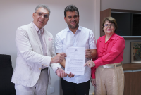 Foto de dois homens altos de roupas claras e uma mulher de calça marrom e camisa rosa. Eles estão em pé posados para a foto segurando um papel branco