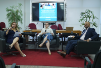 A imagem mostra três pessoas participando de uma roda de conversa em um ambiente formal. Elas estão sentadas em poltronas pretas dispostas sobre um tapete vermelho. A mulher à esquerda, vestindo um vestido azul, segura um microfone e algumas folhas de papel. No centro, uma mulher com camisa clara e saia jeans também segura um microfone, enquanto à direita um homem de terno azul e gravata observa atentamente. 