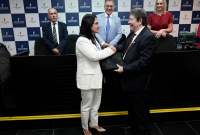 Mulher de terno branco recebendo uma mochila de um homem branco de terno escuro, sendo observado por dois homens e uma mulher