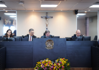 Há uma bancada com três homens sentados. Eles vestem ternos escuros. Também há uma mulher.