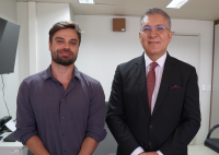Há duas pessoas em pé, lado a lado.  À esquerda, há um homem com barba curta, cabelo castanho e camisa social roxa, com expressão serena. À direita, há um homem de terno preto, camisa branca e gravata bordô, usando óculos. Ele está com as mãos cruzadas à frente do corpo e sorri levemente. 