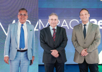 Há três homens posando para foto em um evento, diante de um painel luminoso com palavras parcialmente visíveis ao fundo.  À esquerda, um homem usa terno azul-claro quadriculado, camisa branca e gravata azul. No centro e à direita, dois homens estão lado a lado: o do meio veste terno escuro com camisa clara e gravata listrada em vermelho e azul; o da direita usa terno em tom bege, camisa clara e gravata preta. Todos sorriem e estão em posição formal.