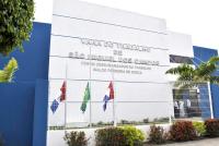 Fachada do prédio daS VaraS do Trabalho de São Miguel dos Campos. A construção é branca e azul, com letras em relevo indicando o nome da unidade. Há uma grade branca na frente, com três bandeiras hasteadas atrás dela. Em primeiro plano, há plantas e arbustos coloridos. À direita, próximo à entrada, algumas pessoas aparecem de pé
