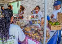 Há uma feira ao ar livre, com uma barraca coberta por lona. Sobre a mesa, há vários produtos artesanais e alimentos embalados — bolos, biscoitos, doces, temperos, geleias e frutas. Três pessoas ficam atrás da bancada atendendo, enquanto outra pessoa, vista de costas, observa os itens. O ambiente é colorido e movimentado, com clima de feira.