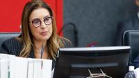 Mulher branca de cabelos loiros usando óculos e sentada; à sua frente, tem um computador preto