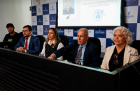 A imagem mostra uma mesa de autoridades durante uma solenidade. Cinco pessoas estão sentadas lado a lado, em um ambiente formal, com painel institucional ao fundo. Ao centro, um homem de terno e gravata fala ou ouve atentamente, ladeado por outros participantes — homens e mulheres — também trajando roupas formais. Um telão ao fundo exibe informações sobre o curso de “Perito Judicial e Assistente Técnico em Radiologia Legal”, indicando o contexto da formatura mencionada no texto.
