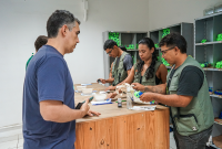 A imagem mostra um grupo de pessoas em um ambiente interno, possivelmente um almoxarifado ou oficina. Quatro pessoas, vestindo coletes verdes, estão em pé atrás de um balcão de madeira manuseando e conferindo kits com frascos e embalagens. À frente, um homem de camiseta azul observa ou aguarda atendimento. Ao fundo, há prateleiras metálicas com vários itens organizados, incluindo objetos de cor verde. A cena transmite a ideia de entrega ou distribuição de materiais.