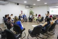 Sala de reuniões bem iluminada, com várias pessoas sentadas em cadeiras organizadas em círculo. A maioria é mulher. Algumas delas seguram cadernos ou laptops. Uma mulher em pé à direita fotografa o encontro com o celular. No centro, um homem de camisa azul fala ao microfone, enquanto os demais ouvem com atenção. Ao fundo, há mesas, uma planta, um quadro branco e um telão