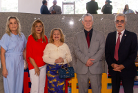 A imagem mostra cinco pessoas posando para uma foto em um ambiente interno bem iluminado. Elas estão lado a lado, sorrindo, em frente a uma parede de granito cinza. As três mulheres à esquerda vestem roupas em tons claros e coloridos — uma em azul, outra em vermelho com calça branca e a terceira com blusa clara e saia estampada. À direita, dois homens usam ternos formais, um em cinza e o outro em preto com gravata vermelha. Ao fundo, é possível ver parte de um espaço infantil, com cercadinho colorido e brin