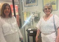 Há duas mulheres vestidasd de branco. Elas estão sorrindo. Logo atrás há um vitrine com fotos e objetos.