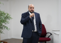 Homem branco calvo de terno escuro e camisa branca. Ele fala ao microfone.