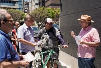Homem de camisa rosa e chapeú bege com uma cartilha na mão passando orientações a ciclistas. Todos estão em pé.