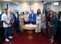 HÁ um grupo de 12 pessoas posando para uma foto oficial em uma sala de reuniões. Elas estão alinhadas atrás de uma mesa redonda de madeira, onde há documentos. O ambiente tem bandeiras ao fundo, um crucifixo na parede e um monitor com horário. As pessoas estão bem-vestidas, algumas de terno e outras com roupas sociais, indicando um evento institucional ou cerimônia formal.