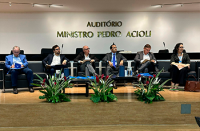 Cinco homens e uma mulher todos sentado no palco de um auditório; todos eles usam ternos escuros, só um homem está usando terno azul claro.