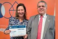 A imagem mostra duas pessoas sorrindo diante de um fundo laranja com elementos gráficos geométricos.  À esquerda, uma mulher segura um certificado de premiação. Ela usa uma blusa estampada em azul e branco, brincos de pérola e um colar discreto. O certificado que ela exibe indica a conquista de 2º lugar na categoria Projeto Gráfico, referente à “Cartilha Diversidade e a Comunidade LGBTQIAPN+”, no Prêmio Justiça do Trabalho de Comunicação Social.  À direita, está um homem vestindo terno cinza, camisa branca.