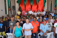 Grupo de pessoas está reunido em um espaço decorado com cortinas vermelhas e verdes e uma árvore de Natal ao fundo. Na primeira fileira, estão homens e mulheres adultos, alguns olhando para a câmera. Ao fundo, também há pessoas posando para a foto. Todos parecem participar de um evento natalino 